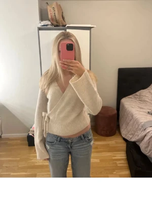 Beige omlottkofta med knytband - Säljer en snygg beige omlottkofta med knytband i sidan och v-ringad hals. Koftan har långa ärmar och är gjord i ett mjukt, stickat material som känns riktigt skönt mot huden. Perfekt att matcha med jeans för en avslappnad look.