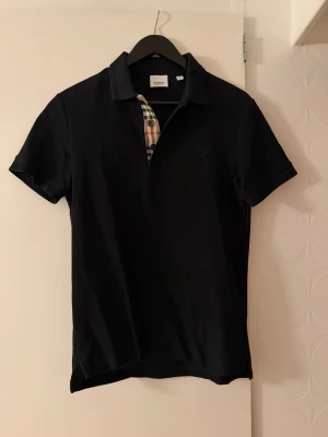 Svart Burberry piké - Klassisk svart pikétröja från Burberry med den ikoniska rutiga detaljen vid knäppningen och diskret broderad logga på bröstet. Tröjan har korta ärmar och mjukt bomullsmaterial som känns skönt mot huden. Perfekt för en stilren och avslappnad look. Köpt på NK, kvitto finns. Använd max 5 gånger.