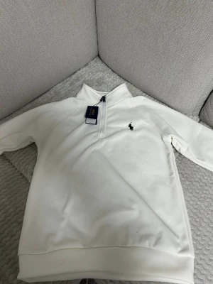 Vit halvzip från Polo Ralph Lauren - Snygg vit tröja med halv dragkedja från Polo Ralph Lauren. Klassisk design med svart broderad logga på bröstet och ribbade muddar. Perfekt för en clean och stilren look.