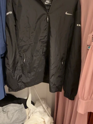 Svart Nike vindjacka med dragkedja - Svart vindjacka från Nike med klassisk logga på bröstet och ärmen. Jackan har hel dragkedja framtill och två fickor med dragkedja. Tillverkad i lätt och smidigt syntetmaterial, perfekt för blåsiga dagar. Enkel och sportig design som funkar till många outfits.