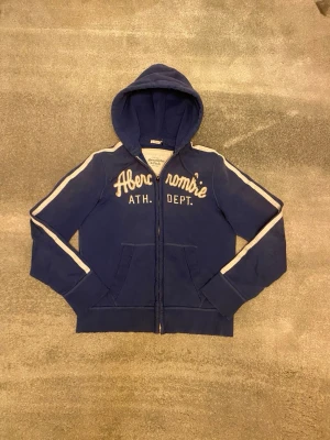 Blå hoodie från Abercrombie - Snygg blå hoodie med dragkedja från Abercrombie & Fitch. Stor vit logga och text på bröstet, vita ränder längs ärmarna och två fickor framtill. Mjuk bomullskänsla och ribbade muddar. Tröjan är i XL men passar nog också L. Vid funderingar, fråga på! 
