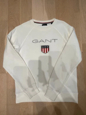 Vit sweatshirt från GANT med tryck - Säljer en vit sweatshirt från GANT med broderat logotyptryck och emblem på bröstet. Tröjan har rund hals, långa ärmar och ribbade muddar. Klassisk och stilren design med grått GANT-tryck och rött, vitt och blått emblem.