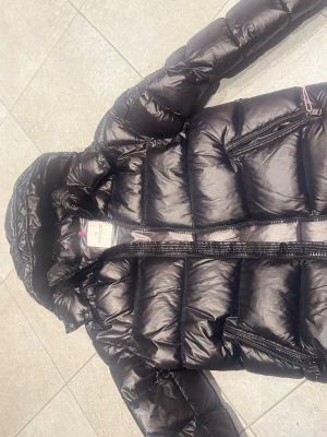 Svart dunjacka från Moncler - Snygg svart dunjacka från Moncler med glansig finish, huva och dragkedja framtill. Jackan har en ficka med lock och Moncler-logga på bröstet. Perfekt för kalla dagar när du vill hålla stilen.