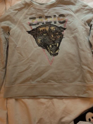 Ljusblå sweatshirt från zadig - Cool ljusblå sweatshirt från Zadig med ett stort pantermotiv i paljetter på bröstet och texten 'ZADIG' ovanför. Tröjan har rund halsringning och långa ärmar. Perfekt för dig som gillar statementplagg och djurmotiv. I storlek 14 år/S