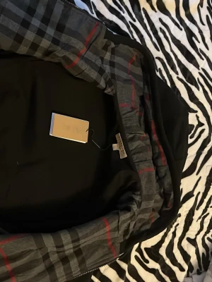 Svart jacka med rutigt foder passar både S och m  - Snygg svart jacka från Burberry med klassiskt rutigt foder i grått, svart och rött. Jackan har dragkedja och en stilren design som passar perfekt till vardagslooken. Märkets ikoniska rutmönster syns tydligt på insidan.