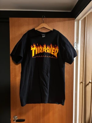 Svart Thrasher t-shirt  - Säljer en svart Thrasher t-shirt med den ikoniska flames-loggan i gult och rött på bröstet. Klassisk passform och rund halsringning. Perfekt för dig som gillar streetwear och skatekultur.