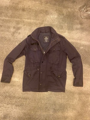 Mörkblå fieldjacka från superdry  - Rikitgt snygg mörkblå fieldjacka från Superdry med fyra framfickor, hög krage och dragkedja samt knappar framtill. Jackan har en klassisk passform och är tillverkad i slitstark material. Vid funderingar, fråga på!