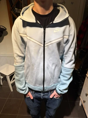 Nike Tech Fleece - Nike Tech Fleece hoodie i ljusblått med dragkedja och vit logga på bröstet. Hoodien har huva, svarta detaljer, fickor på sidorna och en extra ficka med dragkedja på ärmen. Tillverkad i mjukt fleecematerial för maximal komfort.