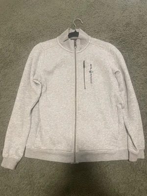 Ljusgrå ziptröja från Sail Racing - Ljusgrå ziptröja från Sail Racing med hög krage och hel dragkedja framtill. Tröjan har en bröstficka med dragkedja och Sail Racing-logga broderad på vänster sida. Tillverkad i mjukt bomullsmaterial och har ribbade muddar vid ärmslut och nederkant.
