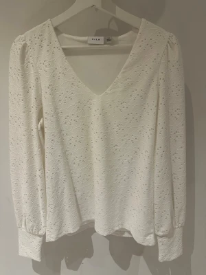Vit spetsblus från Vila storlek S - Söt vit blus från Vila med v-ringning och långärmad design. Blusen har ett broderat spetsmönster med små hål och breda muddar vid ärmsluten. Perfekt för dig som gillar romantisk och feminin stil. Använd endast 1 gång. Längd 58cm