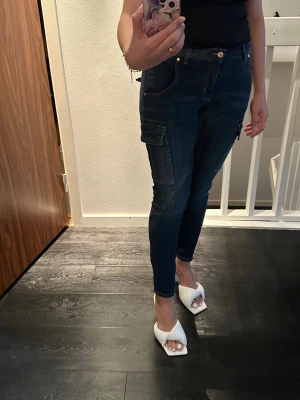 Mörkblå lågmidja  cargopants med fickor - Ljusa jeans med dragkedjedetaljer framtill. I använd skick, en ficka är trasig se bild. De upplevs som 36 i storlek. Jag är 160 cm lång. 