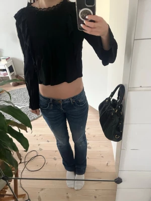 Svart croppad topp med spets från Zara - Trendig svart croppad topp från Zara med lång ärm och snygg spetsdetalj vid halsen. Toppen har en lätt ribbad struktur och en feminin, lite lös passform. 