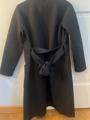 Svart kappa från Pieces med bälte - Stilren svart kappa från Pieces med klassisk krage, stora fickor framtill och ett knytbart bälte i midjan. Kappan har lång ärm och rak passform, perfekt för dig som gillar en enkel men snygg look.