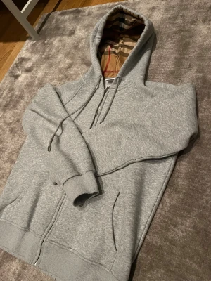 Grå zip hoodie från Burberry - Snygg grå zip hoodie från Burberry med klassiskt rutigt foder i huvan. Hoodien har dragsko i huvan, fickor framtill och ribbade muddar. Perfekt för dig som vill ha en stilren och exklusiv look med en touch av Burberrys ikoniska design. Pris kan diskuteras ‼️.