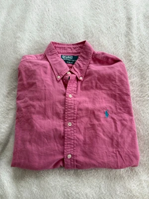Rosa skjorta från Polo Ralph Lauren - Säljer en rosa skjorta från Polo Ralph Lauren i slim fit-modell. Skjortan har klassisk krage, knappar framtill och den ikoniska blå broderade loggan på bröstet. Tillverkad i mjuk bomull och passar perfekt till en stilren look.