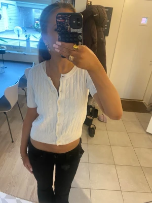 Vit croppad kabelstickad topp - Säljer en vit croppad topp med korta ärmar och kabelstickat mönster. Toppen har knappar framtill och en v-ringad halsringning. Perfekt för dig som gillar en enkel men söt stil. Liten i passform. Har en fläck på insidan som inte går bort, samt en liten sminkfläck på utsidan men inget som syns.