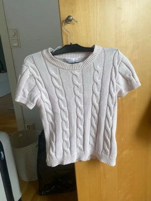 Ljusrosa kabelstickad topp - Säljer en ljusrosa kabelstickad topp med korta ärmar och rund hals. Tröjan har ett klassiskt flätmönster och ribbade kanter vid ärmslut, hals och nederkant. Perfekt för dig som gillar en söt och stilren look.