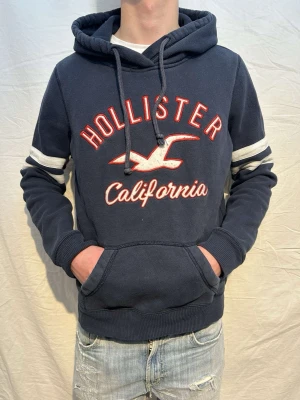 Mörkblå vintage Hollister hoodie - En marinblå vintage hoodie med ett fet tryck!!! Märke:Hollister. Passar perfekt till vintern och våren. Skick 9/10. Size M. Modellen är 176cm och väger 64kg