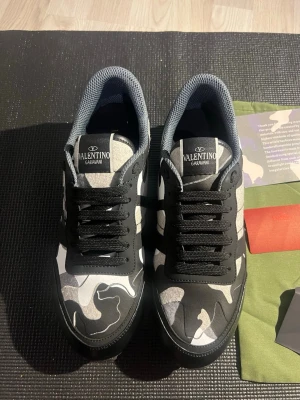 Valentino Garavani camo sneakers - Snygga sneakers från Valentino Garavani med svart och grått camouflagemönster. Skorna har svarta snören, rund tå och platt sula. Märkeslogga på plösen och detaljer i syntetmaterial. Perfekta för dig som vill sticka ut med en cool streetstyle.