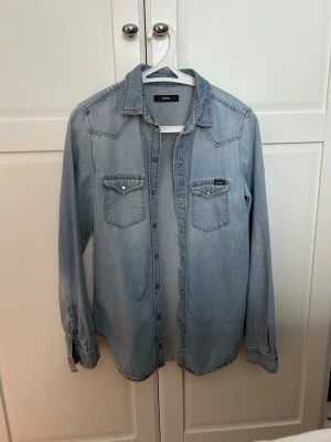 Ljusblå jeansskjorta från Diesel - Säljer en klassisk ljusblå jeansskjorta från Diesel med två bröstfickor och knappar framtill. Skjortan har lång ärm och en avslappnad passform, perfekt att styla lager på lager. Snygga sömdetaljer och ikonisk Diesel-logga på ena fickan. Obetydligt använd, säljs då den inte används. 