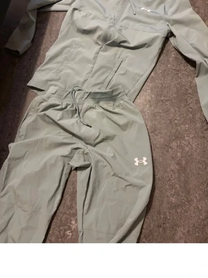 Ljusgrå träningsset från Under Armour - Säljer ett ljusgrått träningsset från Under Armour med dragkedja på jackan och resår i midjan på byxorna. Setet har logga på både jacka och byxa samt texten UNDER ARMOUR på benet. Perfekt för gymmet eller löprundan.