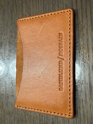 Brun korthållare i skinn Sandlund/Hossain - Slimmad och stilren korthållare i brunt skinn från Sandlund/Hossain. Plånboken har ett minimalistiskt design med ett fack på framsidan och präglad logga. Perfekt för dig som vill ha ett enkelt och snyggt sätt att bära dina kort.