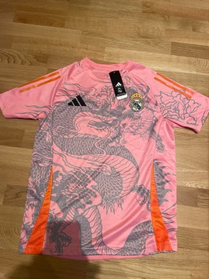 Rosa Real Madrid x Adidas tröja drakmotiv - Snygg rosa sporttröja från Adidas och Real Madrid med grått drakmotiv över hela framsidan. Tröjan har orange detaljer på axlarna och sidorna samt klubbmärke och Adidas-logga på bröstet. Perfekt för dig som vill sticka ut på träningen. Knappt använd