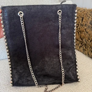 Svart axelväska med kedja och nitar - Cool svart axelväska i mocka med silverfärgade kedjor som axelrem och snygga nitar längs kanterna. Väskan har en fyrkantig form och ger en edgy vibe till din outfit. Perfekt för dig som gillar detaljer och vill sticka ut lite extra.