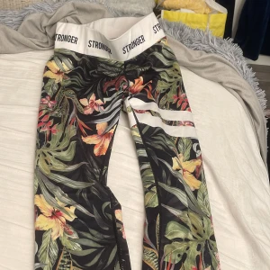 Blommiga leggings från Stronger - Snygga leggings från Stronger med tropiskt blommönster i grönt, gult, orange och rosa. Bred vit resår i midjan med logga och vita ränder på ena benet. Perfekta för träning eller chill.
