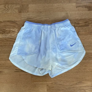 Nike ljusblå träningsshorts - Nike shorts i ljusblått med vitt batikmönster och klassisk Swoosh-logga på ena benet. De har elastisk midja med snörning och är tillverkade i ett lätt, syntetiskt material som andas. Perfekta för löpning eller gymmet, med sportig och avslappnad vibe. Man kan även vika ner toppen biten som på sista bilden för att få en mer lågmidjad look