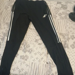 Svarta Adidas träningsbyxor 164 - Svarta träningsbyxor från Adidas med vita ränder längs sidorna och dragkedja vid benslut. Byxorna är tillverkade i återvunnet Primegreen-material och har en smal passform. Perfekta för sport eller chill, med klassisk Adidas-logga på låret.