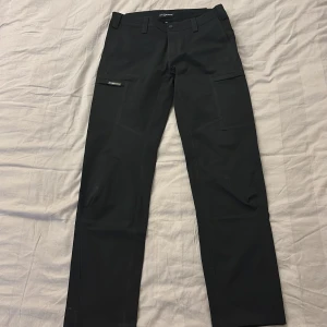 Svarta cargopants från Sail Racing - Svarta cargopants från Sail racing med raka ben, flera praktiska fickor på sidorna och bälteshällor. Byxorna är tillverkade i slitstarkt syntetmaterial och har en stilren look som passar perfekt för uteliv eller vardag. Klassisk passform och diskret logga på fickan.