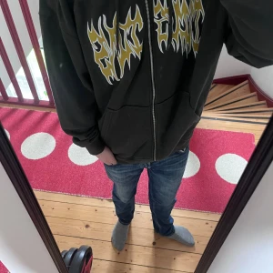 Eftertraktad hoodie med tryck från Weekday - Svart hoodie från Weekday med dragkedja och huva. Framsidan har ett stort, gult och vitt grafiskt tryck i flame-stil. Tröjan har känguruficka och ribbade muddar. Perfekt för dig som gillar streetwear. Storlek S med passform oversized så passar M och L nog ganska bra med.