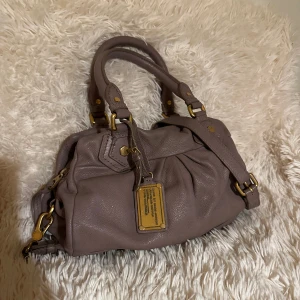 Marc by Marc Jacobs handväska i skinn - Snygg handväska från Marc by Marc Jacobs i en dov lila-brun nyans. Väskan är gjord i mjukt skinn med guldfärgade metalldetaljer och dragkedja. Fodret har coolt logomönster. Perfekt storlek för vardagens essentials och har både handtag och axelrem.