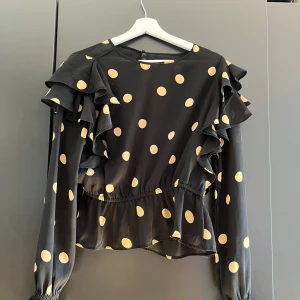 Prickig Svart blus med volanger. - Säljer en svart blus med stora gula prickar och volangdetaljer längs axlarna. Blusen har långärmade ärmar med resår vid handlederna och en lätt rynkad midja som ger en snygg siluett. Materialet är tunt och flowy, perfekt för att sticka ut.