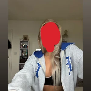 Grå hoodie med blå detaljer - Säljer en grå hoodie med blå huva och blå tryck på bröstet och ärmen. Tröjan har dragkedja framtill och en skön, avslappnad passform. Perfekt för chill dagar eller när du vill ha en sportig vibe.