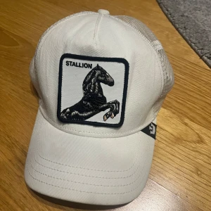 Vit keps Goorin Bros Stallion - Vit keps från Goorin Bros med broderad patch av en svart hingst och texten 'STALLION' framtill. Kepsen har mesh-bakstycke för extra ventilation och justerbar snapback. 