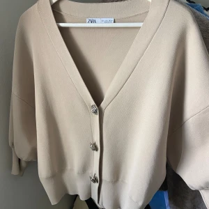 Zara kofta med rosformade knappar - Superfin beige kofta från Zara med v-ringning och tre unika rosformade silvriga knappar framtill. Modellen är lite croppad med breda och puffiga ärmar. Använd fåtal gånger!