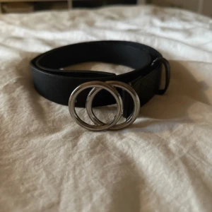 Svart bälte med dubbla ringar - Stilrent svart bälte med två silverfärgade ringar som spänne. Bältet är i skinnimitation och passar perfekt till jeans eller kjol för en trendig look. Enkel design som lyfter outfiten med en cool detalj.