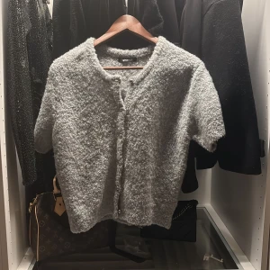 Grå kofta från Gina Tricot - Supermjuk och fluffig grå kofta från Gina Tricot med kort ärm och knappar framtill. Koftan har en rund halsringning och är tillverkad i ett stickat material som känns riktigt cozy. Perfekt att slänga på sig när det är lite kyligt ute.