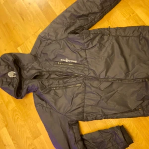 Svart vindjacka från Sail Racing L - Svart vindjacka från Sail Racing med huva och flera dragkedjeförsedda fickor. Jackan är tillverkad i lätt och slitstark polyester, perfekt för blåsiga dagar. Snygg logga på bröstet och innerficka med teknisk info. 100% äkta. Priset kan diskuteras så hör av er. 