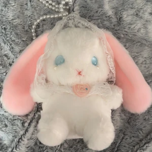 Söt kanin axelväska med pärlband - Supergullig axelväska i form av en kanin med långa rosa öron och vit fluffig päls. Väskan har ett pärlband som axelrem och spetsdetaljer på huvudet. Perfekt för dig som gillar kawaii-stil och vill sticka ut med en unik accessoar.