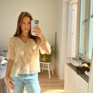 Beige blus med volang och spets - Säljer en superfin beige blus med v-ringning, volangdetaljer och spets längs bröstet. Blusen har korta ärmar, knappar framtill och ett diskret prickigt mönster i tyget. Den är lätt och luftig, perfekt för vår och sommar.