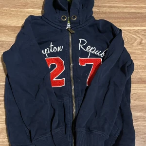 Marinblå hoodie Hampton Republic - Mörkblå hoodie från Hampton Republic med dragkedja, huva och broderad text samt stora röda siffror 27 framtill. Tillverkad i mjuk bomulls- och polyestermix. Perfekt för en avslappnad och sportig look.