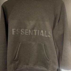 Svart Essentials hoodie med huva - Svart hoodie från Essentials med stor logga framtill och känguruficka. Tröjan har huva och långa ärmar, perfekt för en chill och stilren look. Mjuk insida och klassisk passform.