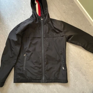 Svart vindjacka från Moncler XL - Svart vindjacka från Moncler med huva och dragkedja framtill. Jackan har broderad Moncler-logga på bröstet, två fickor med dragkedja och snyggt röd, vit och blått band på insidan av huvan. Perfekt för dig som gillar stilren och sportig look.