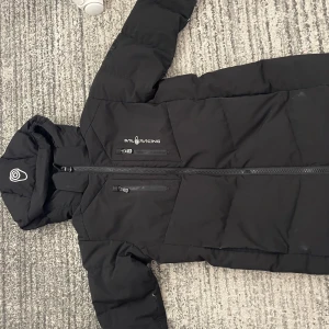 Svart pufferjacka från Sail Racing - Säljer en svart pufferjacka från Sail Racing med huva och dragkedja framtill. Jackan har två bröstfickor med dragkedja och diskret logga på bröstet. Perfekt för kalla dagar och riktigt snygg streetstil.