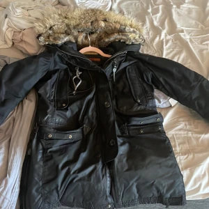 Svart Parajumpers parkas med päls - Svart parkas från Parajumpers med ikonisk patch på ärmen, stor huva med fuskpäls, flera fickor och gul detaljrem bak. Jackan är lång, har dragkedja och knappar framtill samt justerbar midja. Perfekt för kalla dagar.  jackan har ett medel hål i högra fickan