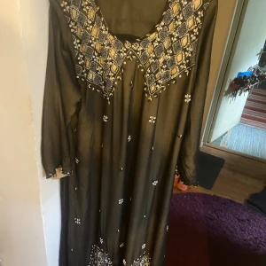 Svart långklänning med broderier - Svart långklänning med vackra broderade detaljer i vitt och guld på bröstet och nertill. Klänningen har lång ärm och ett luftigt, svepande tyg. Perfekt för dig som gillar unika och detaljerade plagg med etnisk touch.