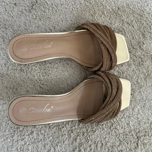 Guldiga sandaler med klack från Cindela - Snygga klackar med låg klack i metallic guld. Skorna har fyrkantig tå och remmar i bronsfärgat material med glittriga detaljer. Perfekta för att lyfta din outfit med en touch av glamour och trendig design.